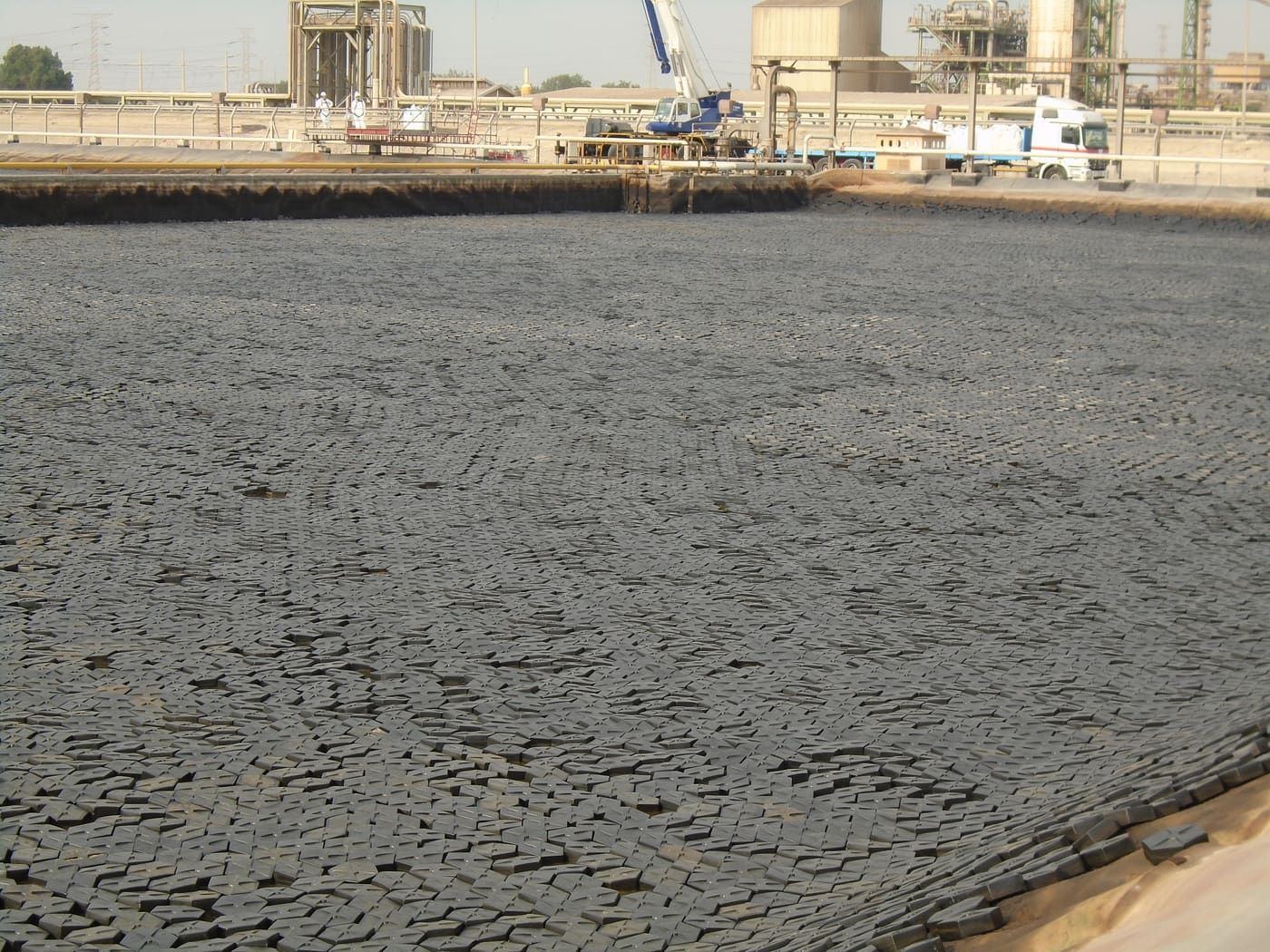An industrial process-water reservoir entirely covered by tightly tessellated black hexagonal Hexprotect AQUA floating tiles, packed edge-to-edge across the whole water surface.