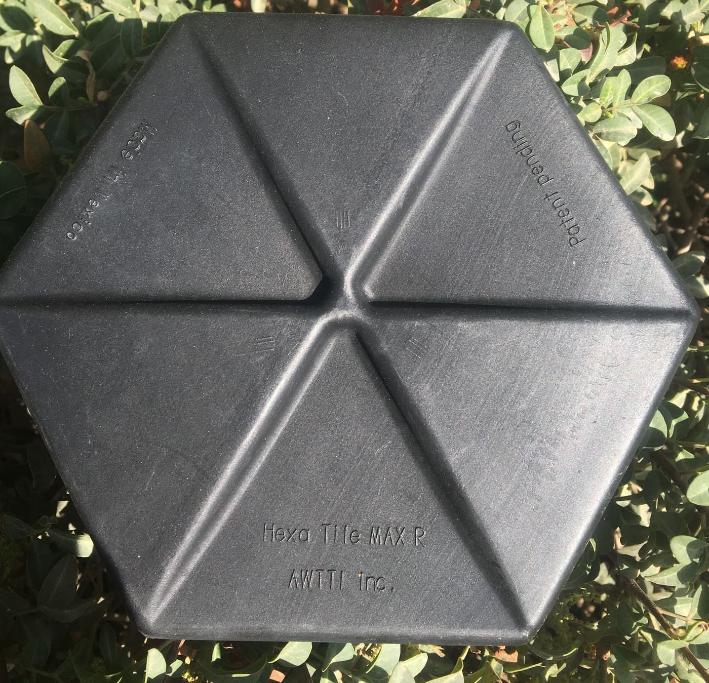 A single black moulded HDPE Hexprotect AQUA hexagonal tile photographed against foliage, showing the six-sided footprint and the raised central dome divided into six triangular facets.